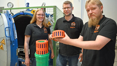 Drei Personen stehen im Hochspannungslabor der FH Dortmund und zeigen ein Bauteil eines Batteriespeichers.