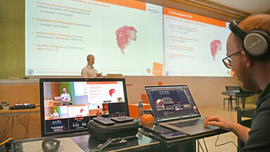 A person is giving a lecture, monitors can be seen in the foreground. The lecture is streamed live online.