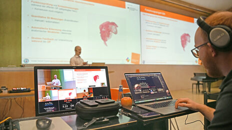 A person is giving a lecture, monitors can be seen in the foreground. The lecture is streamed live online.