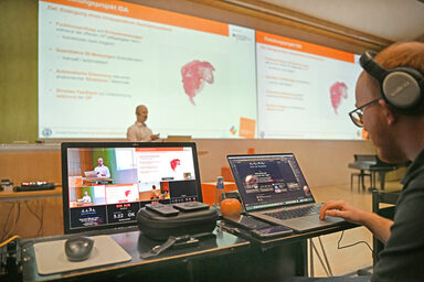 A person is giving a lecture, monitors can be seen in the foreground. The lecture is streamed live online.