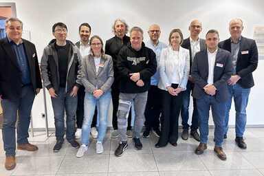 Eleven people stand next to each other in front of a light-colored wall and look friendly into the camera.