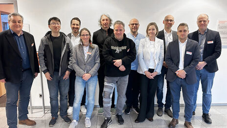 Eleven people stand next to each other in front of a light-colored wall and look friendly into the camera.