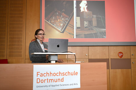 Eine Person links vorne im Bild steht an einem Rednerpult und spricht, hinter ihr sind Bilder eines Geräts zu sehen, auf dem rechten Bild kommen Flammen aus dem Gerät.