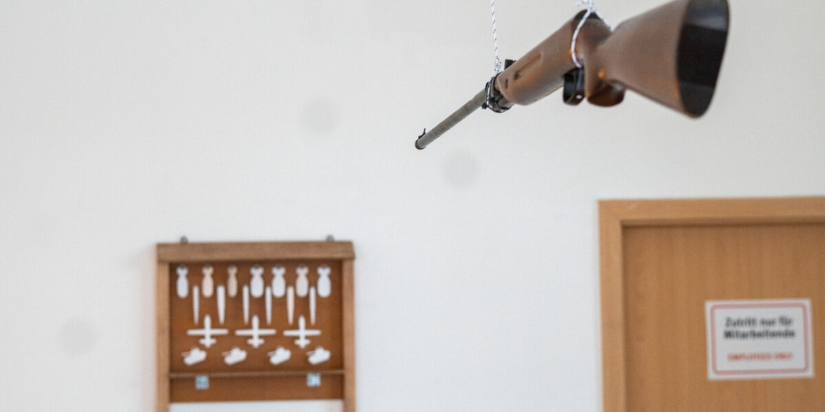 Room installation. A rifle attached to the ceiling is aimed at a small shooting range. The targets are in the shape of bombs and war missiles.