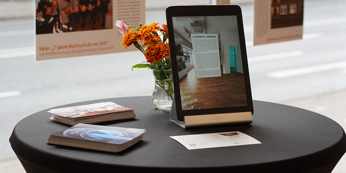 Auf einem Stehtisch befindet sich neben Flyern ein aufgestelltes iPad und eine Vase mit orangenen Blumen