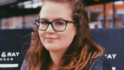 Visiting researcher Dr. Charlotte Rottiers at the Faculty of Architecture. She is wearing a dark sweater and smiling slightly at the camera. Blurred signs and urban structures can be seen in the background.