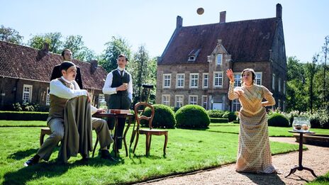 A woman throws a ball in front of a historic building. Three men watch her critically.