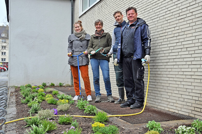 Vier Personen stehen vor einem Gebäude an einem Beet mit Pflanzen, eine Person hält einen Wasserschlauch.