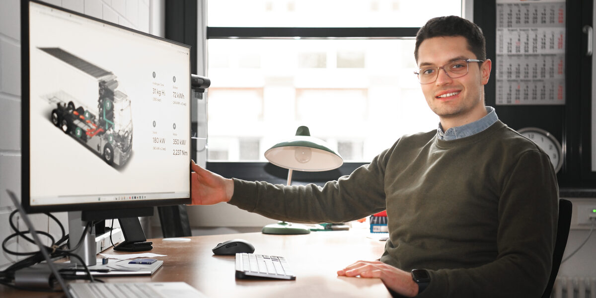 A person sits at a desk and turns the monitor towards the camera, on which a graphic representation of a truck can be seen.