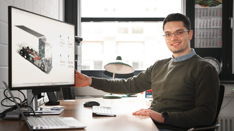 A person sits at a desk and turns the monitor towards the camera, on which a graphic representation of a truck can be seen.