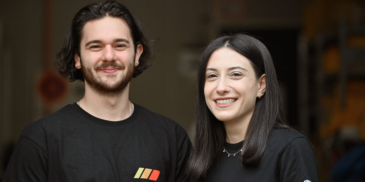 Two people in black T-shirts in a portrait in front of a dark room in the background.