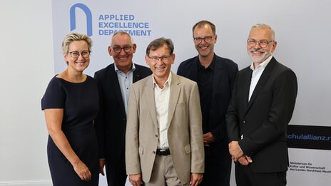 Fünf Personen in repräsentablem Outfit lächeln in die Kamera. Auf dem Banner im Hintergrund ist "Applied Excellence Department" zu lesen.