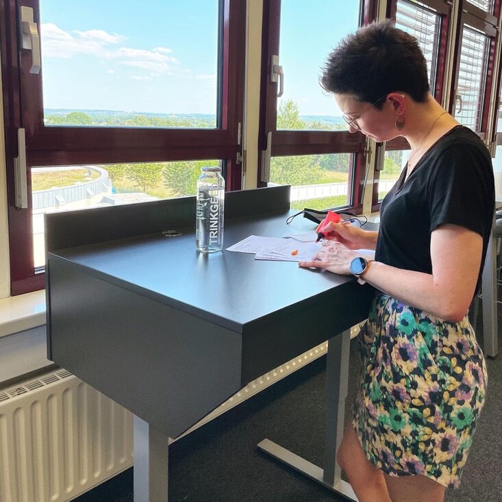 Foto einer Frau, die im Stehen an einem höhenverstellbaren Schreibtisch in der Bibliothek arbeitet__Photo of a woman standing and working on a height-adjustable desk in the library.