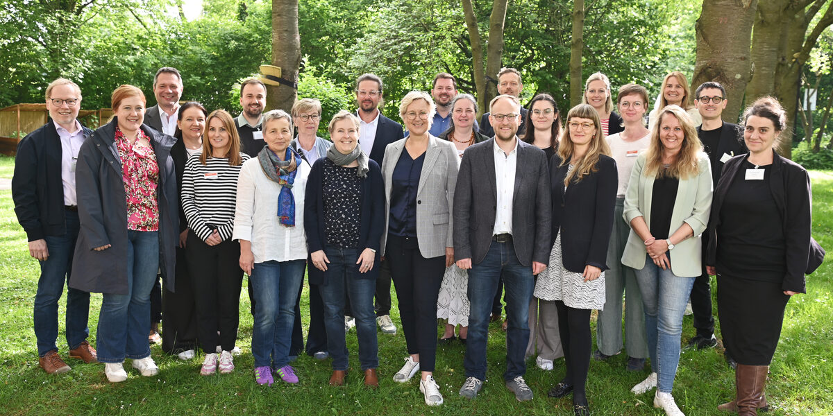 23 Personen stehen auf einer Wiese unter Bäumen und vor Büschen, durch die Baumkronen fällt Sonnenlicht.