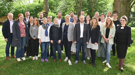 23 people are standing on a meadow under trees and in front of bushes, with sunlight streaming through the treetops.