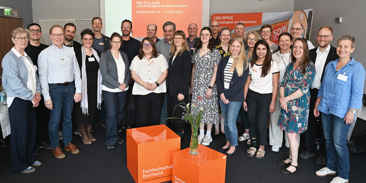 26 people stand in a semi-circle in a room in front of a presentation entitled "HRK pilot Aufdit "Sustainability at universities".