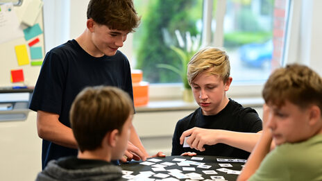 Four students at a high table with small cards representing data. One student holds up a card while the others watch.