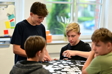 Vier Schüler an einem Stehtisch mit kleinen Karten, die Daten repräsentieren. Ein Schüler hält eine Karte hoch, während die anderen ihm zusehen.