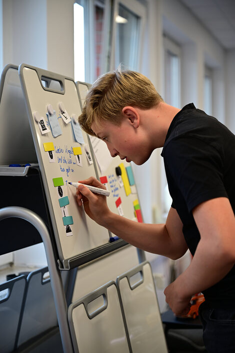 A pupil writes on a whiteboard with a pen. Magnets are used to attach pictograms to the scenario to be worked on.