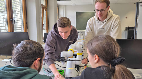 Zwei Personen sitzen an einem Tisch, zwei stehen dahinter. In der Mitte steht ein Roboter mit einem Greifarm.