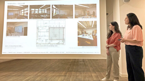 Two female characters stand in front of a screen and present an architectural design.