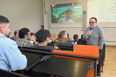 A person read as a male stands in a lecture hall and speaks to a group of students with commitment. Another person is digitally connected on the screen.