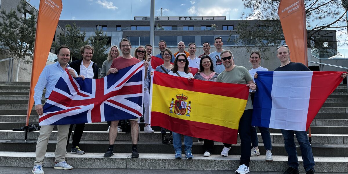 Mehrere Menschen stehen draußen auf einer Treppe, umrahmt von zwei Beachflags der Fachhochschule Dortmund. Die Personen in der ersten Reihe halten vor sich Flaggen der Länder Großbritannien, Spanien und Frankreich.