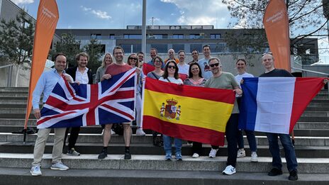 Several people are standing outside on a staircase, framed by two beach flags from Fachhochschule Dortmund. The people in the front row are holding flags of Great Britain, Spain and France in front of them.