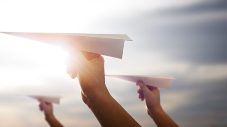 Photo of three hands each holding a paper airplane in front of a sunny sky.