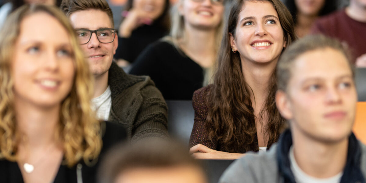 Close-up of several students in the rows of seats in a lecture hall. The focus is on a female student and a male student sitting next to each other and looking straight up.