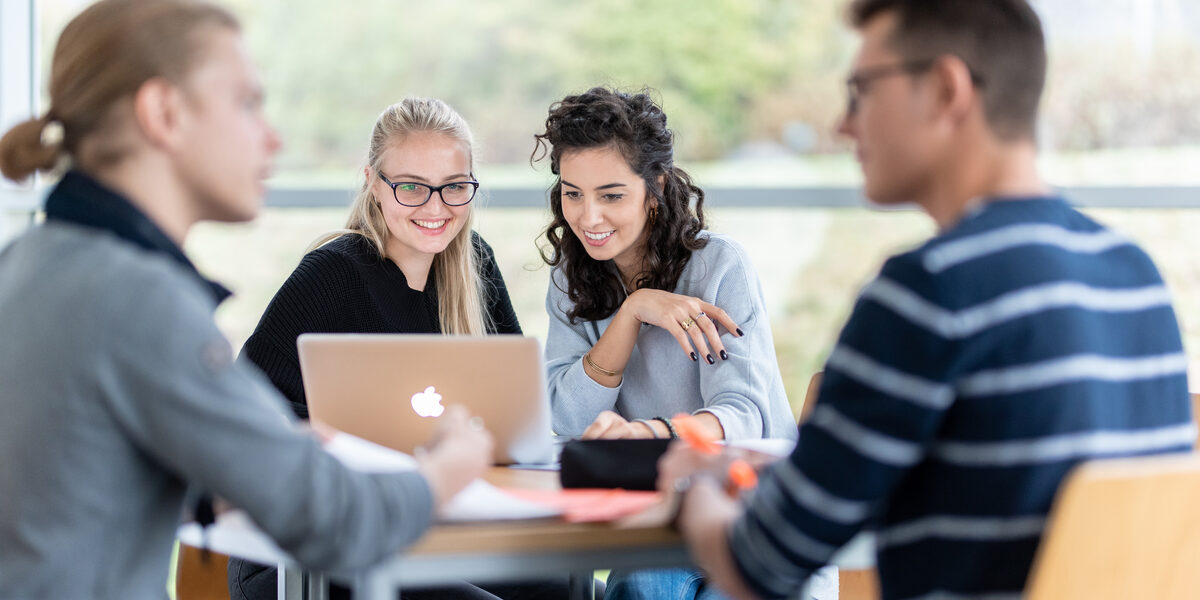 Foto von vier Studierenden an einem Gruppentisch. Im Fokus zwei Studentinnen, die nebeneinander sitzen und auf einen Laptop schauen. Zwei weitere Studenten sind in ein Gespräch vertieft.