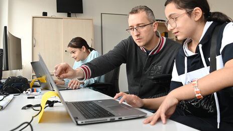 Drei Personen schauen auf Notebook-Monitore. Eine Person erklärt etwas.