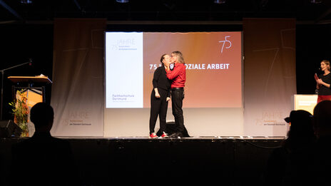 Impressions of the celebrationTwo people on a stage, the one on the right gives the one on the left a kiss on the cheek. A projector image with the text "75 years of social work" can be seen in the background.