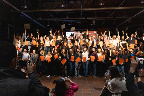 Viele Personen mit orangenen Mappen in der Hand werfen ihre schwarzen Doktorhüte in die Luft, während sie von mehreren Personen im Vordergrund gefilmt werden.