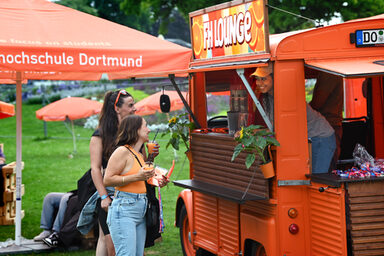 A sign reading "FH Lounge" is attached to the roof of an orange vintage car. From the interior, a person looks outside and smiles at two people standing in front of an open bar in the car, each holding a cup of slush ice, among other things. A meadow with orange parasols can be seen in the background.