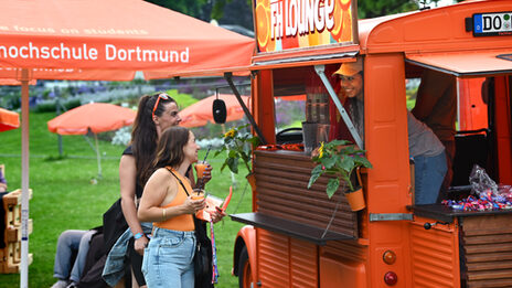 A sign reading "FH Lounge" is attached to the roof of an orange vintage car. From the interior, a person looks outside and smiles at two people standing in front of an open bar in the car, each holding a cup of slush ice, among other things. A meadow with orange parasols can be seen in the background.