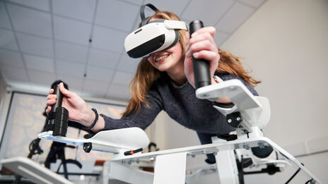 Foto von einer Person auf dem Icaros-Gerät. Sie trägt eine VR-Brille und hält sich an den Griffen fest.
