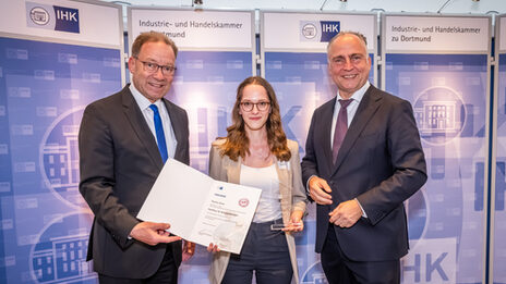 Three people stand next to each other at the presentation of an unfolded certificate and a transparent trophy.