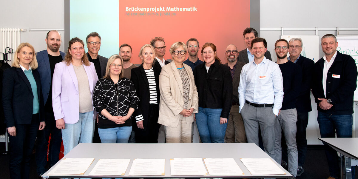 A large group is standing in front of a projector on which the words "Brückenprojekt Mathematik, Feierstunde zum 8. Jubiläum" can be read.