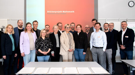 Eine größere Gruppe steht vor einer Beamer-Projektion auf der "Brückenprojekt Mathematik, Feierstunde zum 8. Jubiläum" zu lesen ist.