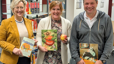 Drei Personen stehen nebeneinander, sie halten jeweils ein Buch vor sich, die Person in der Mitte zusätzlich einen Apfel. Im Hintergrund sind Regale mit weiteren Büchern zu sehen.