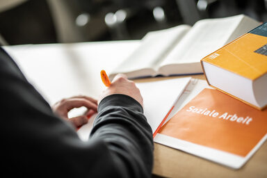 Foto einer Person im Anschnitt, die etwas mit einem orangefarbenen Stift auf einen Block schreibt. Daneben liegen Bücher und ein Heft mit Aufschrift "soziale Arbeit".