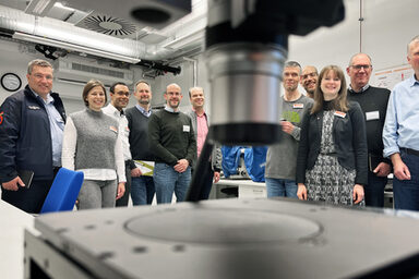 Elf Personen stehen in einem Raum mit dem Gesicht zur Kamera. In der Mitte im Vordergrund ragt von oben, ganz nah und unscharf, ein silberner Zylinder ins Bild. Im unteren Vordergrund, ebenso nah, ist eine dunkle Platte zu sehen.