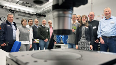 Eleven people stand in a room facing the camera. In the center foreground, a silver cylinder protrudes into the picture from above, very close and out of focus. In the lower foreground, just as close, a dark plate can be seen.
