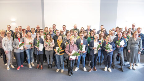 48 people stand in several rows next to each other and look into the camera. Most of them are holding a white envelope and a rose in their hands.