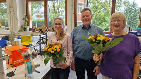 Drei Personen stehen nebeneinander in einem Raum, zwei von ihnen halten jeweils einen Blumenstrauß. Hinter ihnen sind mehrere Gegenstände auf Tischen platziert, unter anderem 3D-Drucker und ein FH-Würfel.