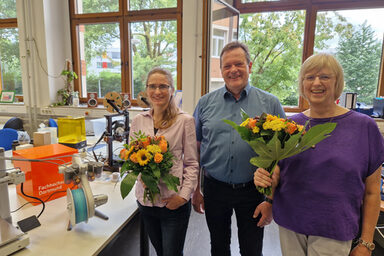 Three people stand next to each other in a room, two of them each holding a bouquet of flowers. Behind them, several objects are placed on tables, including 3D printers and a UAS cube.