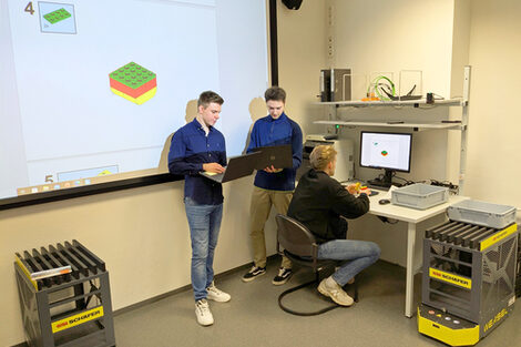 Zwei Personen stehen mit Laptops vor einer Leinwand. Eine dritte Person sitzt vor einem PC an einem Arbeitsplatz.