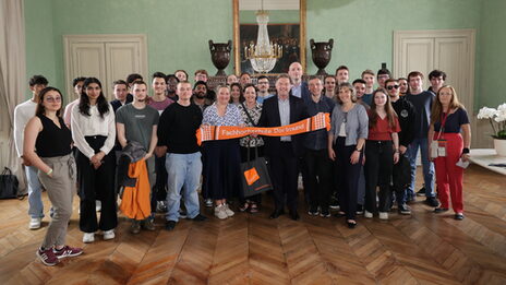 Eine Gruppe von Personen steht in einem Saal, die Personen in der Mitte halten einen orangen Schal mit der Aufschrift Fachhochschule Dortmund.