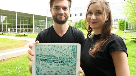 Zwei Personen sitzen auf dem Campus der FH Dortmund. Sie halten ein Tablet in die Kamera. Darauf ist ein architektonischer Entwurf zu sehen.